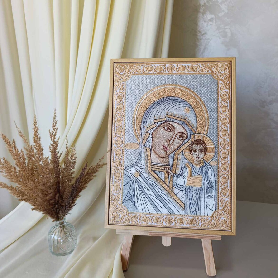 Казанська ікона Божої Матері, вишита шовком, на дерев'яному мольберті поруч зі скляною вазою з сухими колосками