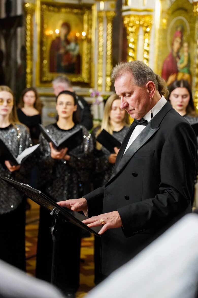 Диригент у чорному фраку перегортає ноти на пюпітрі; позаду хористки в блискучих сріблястих сукнях з нотними папками, на тлі ікон Богородиці в позолочених кіотах