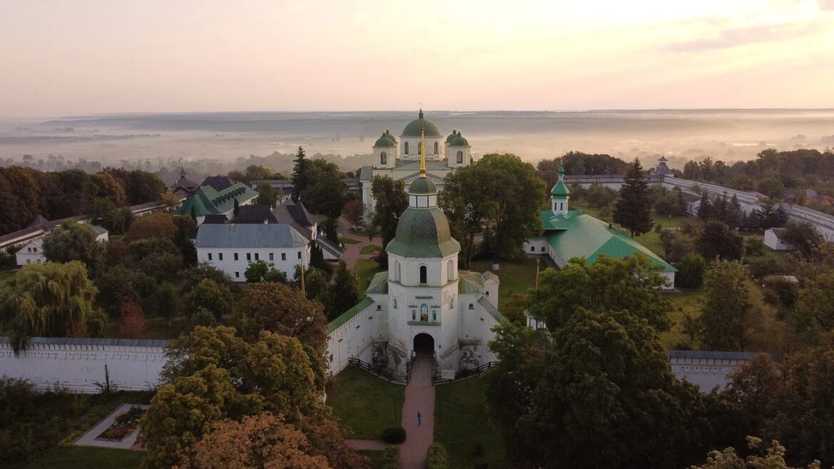 Вигляд згори на білий монастир із зеленими банями: надбрамна дзвіниця з позолоченим шпилем, за нею п'ятибанний собор, довкола мури й дерева, удалині туман над рікою