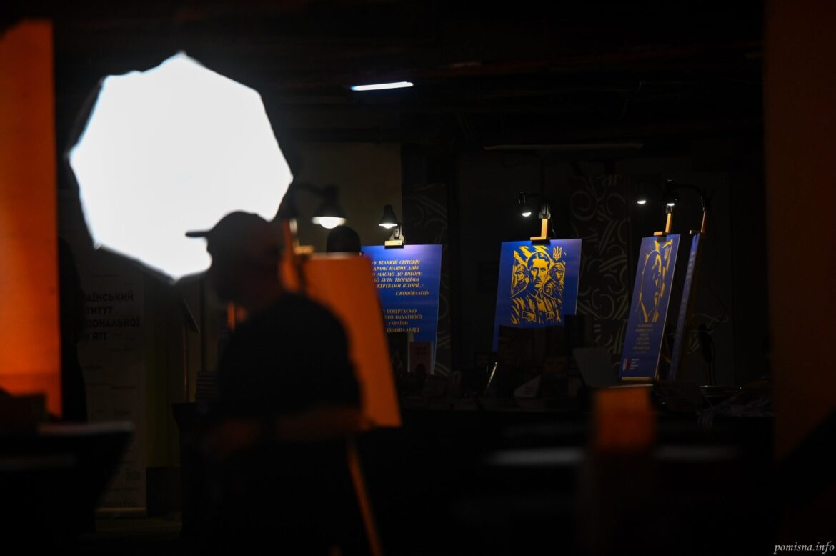 Затемнена зала з виставковими стендами на мольбертах: сині плакати з жовтими портретами й цитатами, підсвічені лампами; на передньому плані силует людини