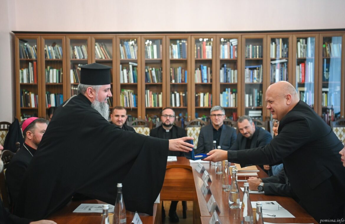 Митрополит Епіфаній через стіл вручає відзнаку в синьому футлярі члену делегації
